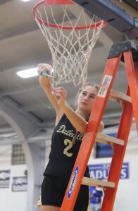 A young female basketball player stands on an orange Werner ladder to cut down the net after a victory. She wears a black Daleville jersey with gold lettering and the number 2 on the front. She holds a pair of scissors in her right hand, carefully snipping a strand of the white mesh net hanging from the basketball hoop. The indoor gymnasium rafters and a blurred background are visible.