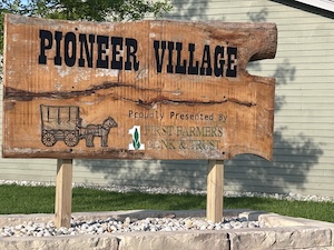 A large, rustic wooden sign with a natural raw edge reads "PIONEER VILLAGE" in bold black, stenciled lettering. Below the text is an illustration of a horse-drawn covered wagon next to a message that reads "Property Presented By First Farmers Bank & Trust" alongside a green and white logo. The sign is mounted on two thick wooden posts rooted inside a raised gravel and stone retaining bed, situated outdoors in front of a light-colored siding building.