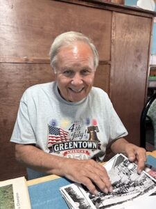 An older man with gray hair and a warm smile sits at a table, looking directly at the camera. He wears a light gray t-shirt featuring a "Happy 4th of July! GREENTOWN" graphic with an American flag, fireworks, and the Liberty Bell. His hands rest on a stack of black-and-white historical photographs spread across the table in front of a large wooden cabinet background.