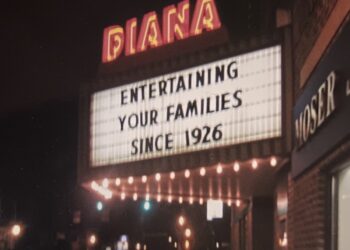 A nighttime view of the glowing neon marquee of the historic Diana Theatre. The theater name "DIANA" is brightly illuminated in red and orange neon lettering above a large, backlit white marquee board. The marquee displays the text "ENTERTAINING YOUR FAMILIES SINCE 1926" in bold black letters. A row of warm incandescent bulbs glows underneath the marquee, casting light onto a dark street setting in front of an adjacent brick building.