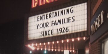 A nighttime view of the glowing neon marquee of the historic Diana Theatre. The theater name "DIANA" is brightly illuminated in red and orange neon lettering above a large, backlit white marquee board. The marquee displays the text "ENTERTAINING YOUR FAMILIES SINCE 1926" in bold black letters. A row of warm incandescent bulbs glows underneath the marquee, casting light onto a dark street setting in front of an adjacent brick building.