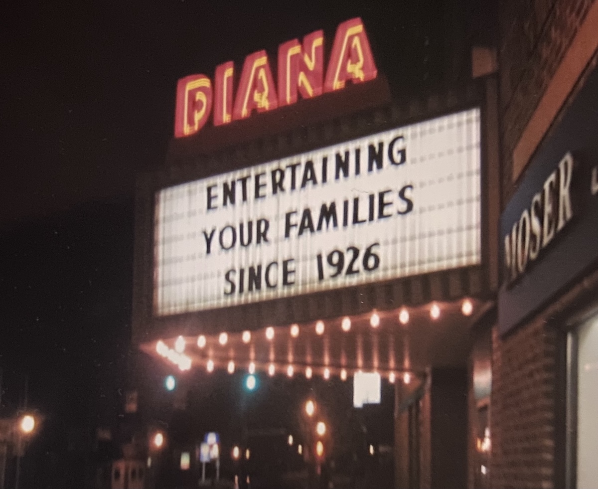 A nighttime view of the glowing neon marquee of the historic Diana Theatre. The theater name "DIANA" is brightly illuminated in red and orange neon lettering above a large, backlit white marquee board. The marquee displays the text "ENTERTAINING YOUR FAMILIES SINCE 1926" in bold black letters. A row of warm incandescent bulbs glows underneath the marquee, casting light onto a dark street setting in front of an adjacent brick building.