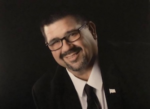A portrait of a smiling man with glasses, short dark hair, and a goatee, wearing a black suit jacket, a white dress shirt, and a black tie, posing against a solid black background.