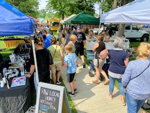 A crowded outdoor market scene filled with people walking along a concrete sidewalk lined with colorful vendor tents on a sunny day. On the left under a blue canopy, a table displays various products, and an A-frame chalkboard sign in the foreground reads "RAW LOCAL HONEY". Green and white vendor tents line the opposite side of the path, where a white vehicle is parked on a grassy area under large trees.
