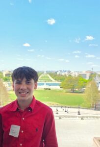 A vertical medium close-up portrait of a smiling young man looking towards the camera, standing on an elevated balcony or terrace overlooking the National Mall in Washington, D.C. He is wearing the same vibrant red long-sleeved button-up shirt with a small white name tag pinned to his chest. In the background, a sprawling panoramic view extends down the green lawns of the National Mall under a bright blue sky with sparse clouds. The Capitol Reflection Pool and distant monuments are visible framed by spring green trees and city buildings.
