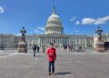 A full-length, wide outdoor portrait of a smiling young person standing on the wide stone plaza directly in front of the United States Capitol building in Washington, D.C. The individual wears a vibrant red long-sleeved button-up shirt, blue jeans, and brown leather dress shoes while giving a thumbs-up gesture. In the background, the grand neoclassical Capitol architecture stands prominently beneath a bright blue sky dotted with scattered white clouds. Two ornate, historical stone lampposts flank the midground, where various tourists, pedestrians, and parked bicycles are visible walking around the plaza.
