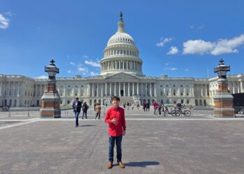 A full-length, wide outdoor portrait of a smiling young person standing on the wide stone plaza directly in front of the United States Capitol building in Washington, D.C. The individual wears a vibrant red long-sleeved button-up shirt, blue jeans, and brown leather dress shoes while giving a thumbs-up gesture. In the background, the grand neoclassical Capitol architecture stands prominently beneath a bright blue sky dotted with scattered white clouds. Two ornate, historical stone lampposts flank the midground, where various tourists, pedestrians, and parked bicycles are visible walking around the plaza.