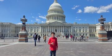 A full-length, wide outdoor portrait of a smiling young person standing on the wide stone plaza directly in front of the United States Capitol building in Washington, D.C. The individual wears a vibrant red long-sleeved button-up shirt, blue jeans, and brown leather dress shoes while giving a thumbs-up gesture. In the background, the grand neoclassical Capitol architecture stands prominently beneath a bright blue sky dotted with scattered white clouds. Two ornate, historical stone lampposts flank the midground, where various tourists, pedestrians, and parked bicycles are visible walking around the plaza.