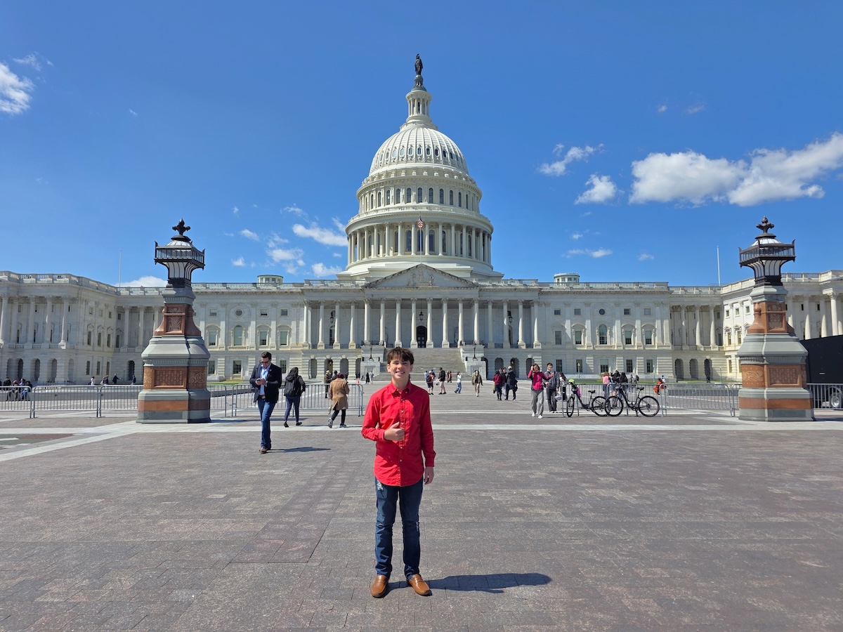 A full-length, wide outdoor portrait of a smiling young person standing on the wide stone plaza directly in front of the United States Capitol building in Washington, D.C. The individual wears a vibrant red long-sleeved button-up shirt, blue jeans, and brown leather dress shoes while giving a thumbs-up gesture. In the background, the grand neoclassical Capitol architecture stands prominently beneath a bright blue sky dotted with scattered white clouds. Two ornate, historical stone lampposts flank the midground, where various tourists, pedestrians, and parked bicycles are visible walking around the plaza.