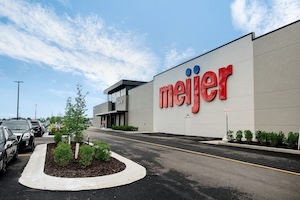 A wide outdoor shot showcases the modern exterior façade of the newly opened Meijer Supercenter in Brownsburg, Indiana. The large white-paneled building features the iconic red "meijer" logo with its two blue dots towering over a freshly paved asphalt drive and parking lot area. A concrete-curbed landscaped island with small green bushes and young trees sits in the foreground on the left, under a bright blue sky filled with wispy white clouds.