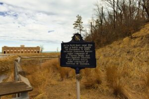 A dark blue historical marker stands in a sandy, grassy dune landscape next to a wooden boardwalk railing. The sign text describes the Battle of Le Petit Fort, fought on December 5, 1780. In the background, a historic brick building with arched openings sits near a body of water under a partly cloudy sky.