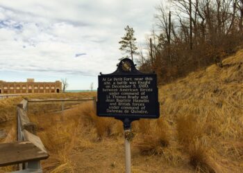A dark blue historical marker stands in a sandy, grassy dune landscape next to a wooden boardwalk railing. The sign text describes the Battle of Le Petit Fort, fought on December 5, 1780. In the background, a historic brick building with arched openings sits near a body of water under a partly cloudy sky.