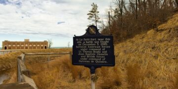 A dark blue historical marker stands in a sandy, grassy dune landscape next to a wooden boardwalk railing. The sign text describes the Battle of Le Petit Fort, fought on December 5, 1780. In the background, a historic brick building with arched openings sits near a body of water under a partly cloudy sky.