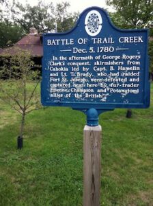 A close-up shot of a blue Battle of Trail Creek Historical Marker with white lettering. The text on the sign describes a Revolutionary War skirmish on Dec. 5, 1780 involving skirmishers from Cahokia, Captain B. Hamelin, and Lieutenant T. Brady. The sign stands on a wooden post in front of a green lawn and a building with a dark, low-slung roof.
