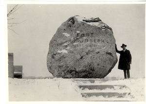 An archival black and white photograph depicting a man in a trench coat and fedora hat standing in the snow next to a massive glacial boulder marker. The large rock has text engraved on its side that reads "FORT ST. JOSEPH". A snowy steps structure sits in the foreground