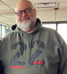 A portrait of a smiling older man with a white beard and glasses, wearing an olive green pullover hoodie featuring a distressed black number 22 graphic with the red text "Veteran Lives Matter" printed across the front chest.