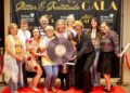Nine people smile cheerfully while posing together on a red carpet at an indoor evening event. They stand before a large black step-and-repeat backdrop decorated with gold lettering that reads "Glitter & Gratitude GALA" and white logos for "Modern Woodmen Fraternal Financial". The attendees are dressed in a mix of colorful, patterned, and sequined outfits, with one individual wearing a bright blonde wig, retro glasses, and a shimmering black button-down shirt. Several people in the middle hold a large, round prop styled to look like a purple vinyl record, covered in signatures with a central label that reads "A Night of Soul and Service – Gala 2026."