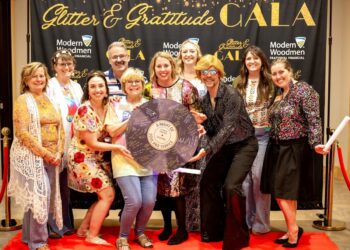 Nine people smile cheerfully while posing together on a red carpet at an indoor evening event. They stand before a large black step-and-repeat backdrop decorated with gold lettering that reads "Glitter & Gratitude GALA" and white logos for "Modern Woodmen Fraternal Financial". The attendees are dressed in a mix of colorful, patterned, and sequined outfits, with one individual wearing a bright blonde wig, retro glasses, and a shimmering black button-down shirt. Several people in the middle hold a large, round prop styled to look like a purple vinyl record, covered in signatures with a central label that reads "A Night of Soul and Service – Gala 2026."