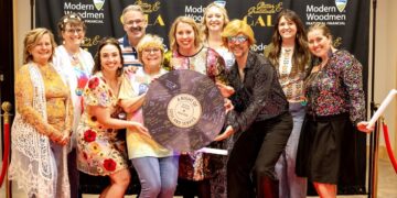 Nine people smile cheerfully while posing together on a red carpet at an indoor evening event. They stand before a large black step-and-repeat backdrop decorated with gold lettering that reads "Glitter & Gratitude GALA" and white logos for "Modern Woodmen Fraternal Financial". The attendees are dressed in a mix of colorful, patterned, and sequined outfits, with one individual wearing a bright blonde wig, retro glasses, and a shimmering black button-down shirt. Several people in the middle hold a large, round prop styled to look like a purple vinyl record, covered in signatures with a central label that reads "A Night of Soul and Service – Gala 2026."