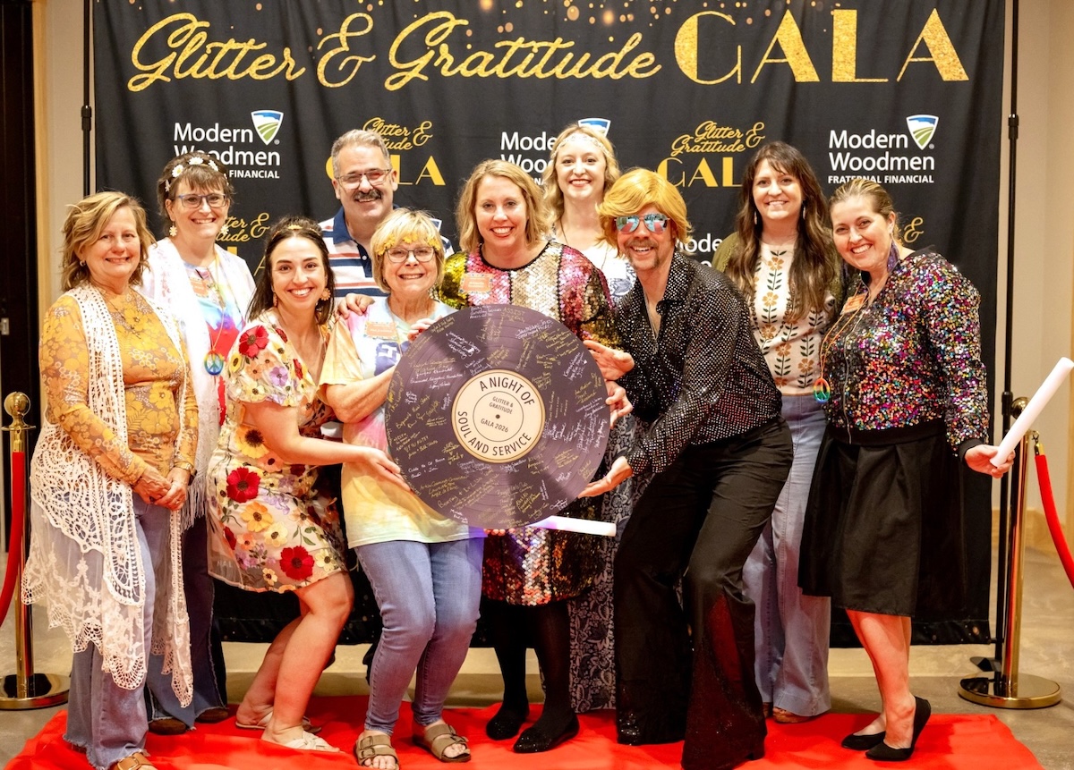 Nine people smile cheerfully while posing together on a red carpet at an indoor evening event. They stand before a large black step-and-repeat backdrop decorated with gold lettering that reads "Glitter & Gratitude GALA" and white logos for "Modern Woodmen Fraternal Financial". The attendees are dressed in a mix of colorful, patterned, and sequined outfits, with one individual wearing a bright blonde wig, retro glasses, and a shimmering black button-down shirt. Several people in the middle hold a large, round prop styled to look like a purple vinyl record, covered in signatures with a central label that reads "A Night of Soul and Service – Gala 2026."