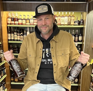 A smiling man with a light beard wearing a black and white trucker hat, a black hoodie, and an unbuttoned tan canvas shirt. He is sitting down and holding two different bottles of bourbon whiskey in his hands, with well-stocked liquor store shelves filled with various spirit bottles blurring out in the background.