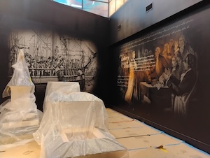 An indoor photograph shows a museum exhibition space under construction or preparation, with the floor protected by paper or cardboard sheets. To the left, several structures or display cases are covered in protective translucent plastic wrapping. The walls are black and feature large historical murals; the left wall depicts a dense crowd of people, while the right wall displays a classic painted scene of historical figures gathered around a table with a white text excerpt overlaid on the imagery.