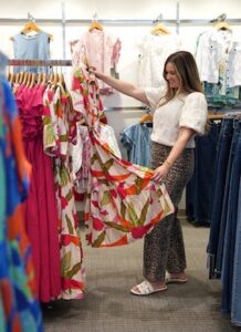 Interior shot of a brightly lit clothing boutique. A woman stands in the center, smiling as she examines a tiered maxi dress with a bold pink and green tropical pattern. A clothing rack to her left features matching tropical dresses and a solid magenta dress. The woman wears a textured, cream-colored short-sleeve top with puffed sleeves, wide-leg leopard print pants, and white slide sandals. Racks of denim and pastel shirts fill the background.
