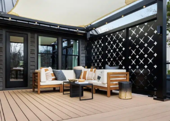 An outdoor composite deck features a modern sectional sofa with wooden frames and cream-colored cushions, adorned with striped and solid accent pillows. The seating area is arranged around a low black coffee table underneath a beige shade sail structure supported by a black frame lined with string lights. A decorative black metal privacy screen with geometric crosscut patterns stands behind the sofa, contrasting against the dark siding of the house.