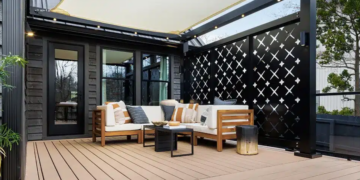 An outdoor composite deck features a modern sectional sofa with wooden frames and cream-colored cushions, adorned with striped and solid accent pillows. The seating area is arranged around a low black coffee table underneath a beige shade sail structure supported by a black frame lined with string lights. A decorative black metal privacy screen with geometric crosscut patterns stands behind the sofa, contrasting against the dark siding of the house.