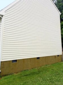 The exterior side wall of a two-story residential house after being cleaned. The light yellow vinyl siding is completely free of mold and algae, appearing bright and clean. The concrete foundation wall below shows some remaining brownish discoloration but is clear of green growth.