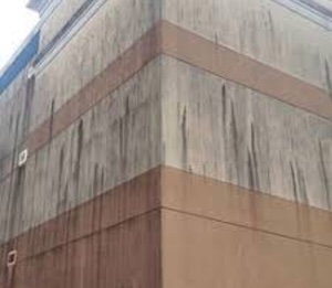 The exterior corner stucco wall of a multi-story commercial building. Heavy, dark black and gray water-dripping stains, mold, and environmental pollution grime cover the beige and brown painted sections of the building facade.