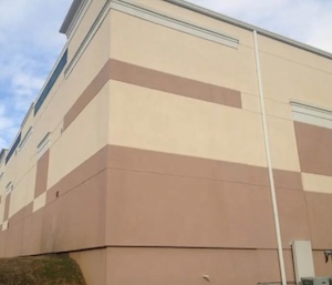 The exterior corner stucco wall of a multi-story commercial building after being cleaned. The beige and brown painted sections of the facade are entirely free of black water stains, mold, and environmental grime, appearing clean and uniform under a partly cloudy sky.