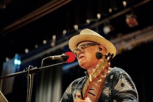 A low-angle, close-up shot of a male musician singing into a microphone on a stage. He is wearing a straw fedora hat, glasses, an in-ear monitor, and a blue and grey patterned button-down shirt while playing an acoustic guitar. The stage background is dark with warm, dramatic theatrical lighting and equipment rigging visible overhead.