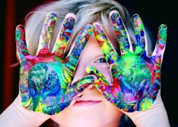 A close-up portrait of a young child with blonde hair peeking through their hands, which are held up in front of their face. The child's palms and fingers are completely covered in thick, swirled layers of vibrant, multicolored acrylic paint in shades of green, blue, yellow, and red. One blue eye is visible through a small gap between their fingers, and they have a soft, subtle smile against a dark background.