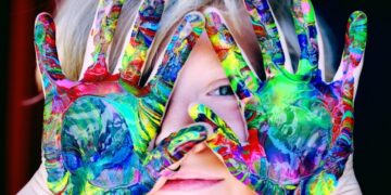 A close-up portrait of a young child with blonde hair peeking through their hands, which are held up in front of their face. The child's palms and fingers are completely covered in thick, swirled layers of vibrant, multicolored acrylic paint in shades of green, blue, yellow, and red. One blue eye is visible through a small gap between their fingers, and they have a soft, subtle smile against a dark background.