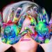 A close-up portrait of a young child with blonde hair peeking through their hands, which are held up in front of their face. The child's palms and fingers are completely covered in thick, swirled layers of vibrant, multicolored acrylic paint in shades of green, blue, yellow, and red. One blue eye is visible through a small gap between their fingers, and they have a soft, subtle smile against a dark background.