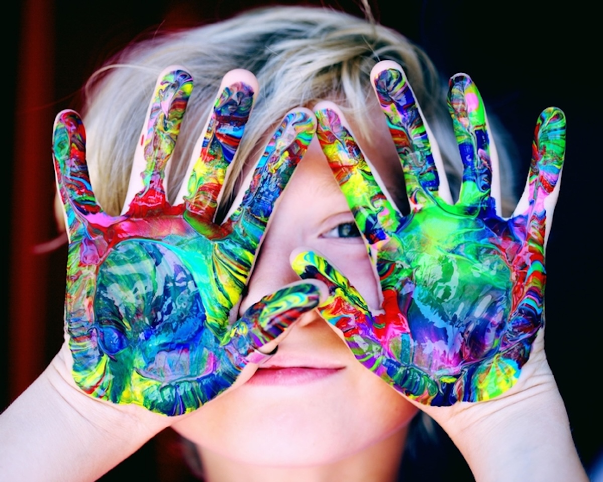 A close-up portrait of a young child with blonde hair peeking through their hands, which are held up in front of their face. The child's palms and fingers are completely covered in thick, swirled layers of vibrant, multicolored acrylic paint in shades of green, blue, yellow, and red. One blue eye is visible through a small gap between their fingers, and they have a soft, subtle smile against a dark background.