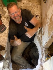 High-angle view of a smiling man with safety glasses and a goatee sitting inside a large trench cut out of a tiled floor. He is wearing a black t-shirt and work pants, giving a double thumbs-up while working around underground plumbing pipes.
