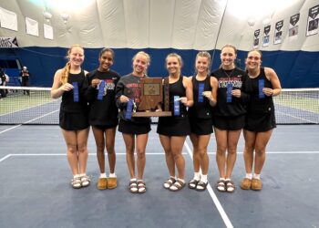 Seven female athletes from the Center Grove Trojans tennis team stand shoulder-to-shoulder on a blue indoor tennis court. They are dressed in matching black athletic uniforms, with several wearing black sweatshirts that read "Center Grove Tennis." In the center, two players hold a large wooden IHSAA championship trophy cut into the shape of Indiana. Most players hold up blue award ribbons, and all are smiling broadly for the camera. A tennis net stretches behind them inside a large dome facility.