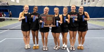 Seven female athletes from the Center Grove Trojans tennis team stand shoulder-to-shoulder on a blue indoor tennis court. They are dressed in matching black athletic uniforms, with several wearing black sweatshirts that read "Center Grove Tennis." In the center, two players hold a large wooden IHSAA championship trophy cut into the shape of Indiana. Most players hold up blue award ribbons, and all are smiling broadly for the camera. A tennis net stretches behind them inside a large dome facility.