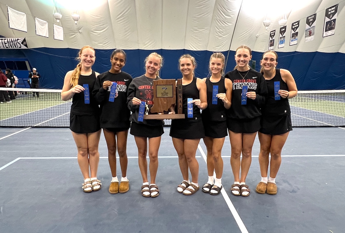 Seven female athletes from the Center Grove Trojans tennis team stand shoulder-to-shoulder on a blue indoor tennis court. They are dressed in matching black athletic uniforms, with several wearing black sweatshirts that read "Center Grove Tennis." In the center, two players hold a large wooden IHSAA championship trophy cut into the shape of Indiana. Most players hold up blue award ribbons, and all are smiling broadly for the camera. A tennis net stretches behind them inside a large dome facility.