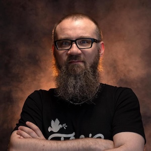 A portrait of a man with black-rimmed glasses and a long, full beard posing with his arms crossed. He is wearing a black t-shirt featuring a white dove graphic against a dark brown, textured backdrop with rim lighting on his hair and shoulders.