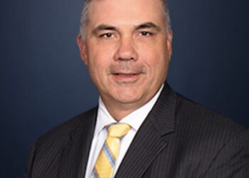 A formal professional headshot of a middle-aged man with short graying hair, wearing a dark pinstripe suit jacket, a white dress shirt, and a yellow, blue, and white striped tie, smiling slightly against a dark blue background.