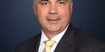 A formal professional headshot of a middle-aged man with short graying hair, wearing a dark pinstripe suit jacket, a white dress shirt, and a yellow, blue, and white striped tie, smiling slightly against a dark blue background.