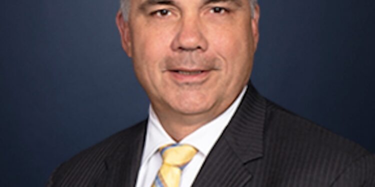 A formal professional headshot of a middle-aged man with short graying hair, wearing a dark pinstripe suit jacket, a white dress shirt, and a yellow, blue, and white striped tie, smiling slightly against a dark blue background.