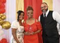 A smiling group of three people pose for a formal award photo in front of a decorative backdrop featuring a red curtain flanked by shimmering gold sequin drapes. On the left, a younger woman with styled black hair wearing a white dress with a floral pattern smiles warmly. In the center, an older woman with an elegant braided updo wears an off-the-shoulder red dress and proudly holds a clear acrylic shield award that reads "Roses and Pearls 2026 Rosa Legacy Award - Carolyn Reeves". On the right, a bearded man wearing glasses, a black vest, a white shirt, and a black tie stands with his hand in his pocket. The scene is framed by a white and gold balloon arch on the far left.