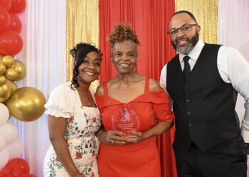 A smiling group of three people pose for a formal award photo in front of a decorative backdrop featuring a red curtain flanked by shimmering gold sequin drapes. On the left, a younger woman with styled black hair wearing a white dress with a floral pattern smiles warmly. In the center, an older woman with an elegant braided updo wears an off-the-shoulder red dress and proudly holds a clear acrylic shield award that reads "Roses and Pearls 2026 Rosa Legacy Award - Carolyn Reeves". On the right, a bearded man wearing glasses, a black vest, a white shirt, and a black tie stands with his hand in his pocket. The scene is framed by a white and gold balloon arch on the far left.