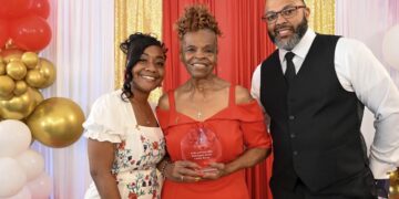 A smiling group of three people pose for a formal award photo in front of a decorative backdrop featuring a red curtain flanked by shimmering gold sequin drapes. On the left, a younger woman with styled black hair wearing a white dress with a floral pattern smiles warmly. In the center, an older woman with an elegant braided updo wears an off-the-shoulder red dress and proudly holds a clear acrylic shield award that reads "Roses and Pearls 2026 Rosa Legacy Award - Carolyn Reeves". On the right, a bearded man wearing glasses, a black vest, a white shirt, and a black tie stands with his hand in his pocket. The scene is framed by a white and gold balloon arch on the far left.