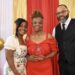 A smiling group of three people pose for a formal award photo in front of a decorative backdrop featuring a red curtain flanked by shimmering gold sequin drapes. On the left, a younger woman with styled black hair wearing a white dress with a floral pattern smiles warmly. In the center, an older woman with an elegant braided updo wears an off-the-shoulder red dress and proudly holds a clear acrylic shield award that reads "Roses and Pearls 2026 Rosa Legacy Award - Carolyn Reeves". On the right, a bearded man wearing glasses, a black vest, a white shirt, and a black tie stands with his hand in his pocket. The scene is framed by a white and gold balloon arch on the far left.