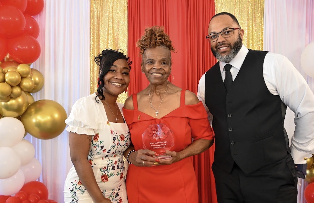 A smiling group of three people pose for a formal award photo in front of a decorative backdrop featuring a red curtain flanked by shimmering gold sequin drapes. On the left, a younger woman with styled black hair wearing a white dress with a floral pattern smiles warmly. In the center, an older woman with an elegant braided updo wears an off-the-shoulder red dress and proudly holds a clear acrylic shield award that reads "Roses and Pearls 2026 Rosa Legacy Award - Carolyn Reeves". On the right, a bearded man wearing glasses, a black vest, a white shirt, and a black tie stands with his hand in his pocket. The scene is framed by a white and gold balloon arch on the far left.